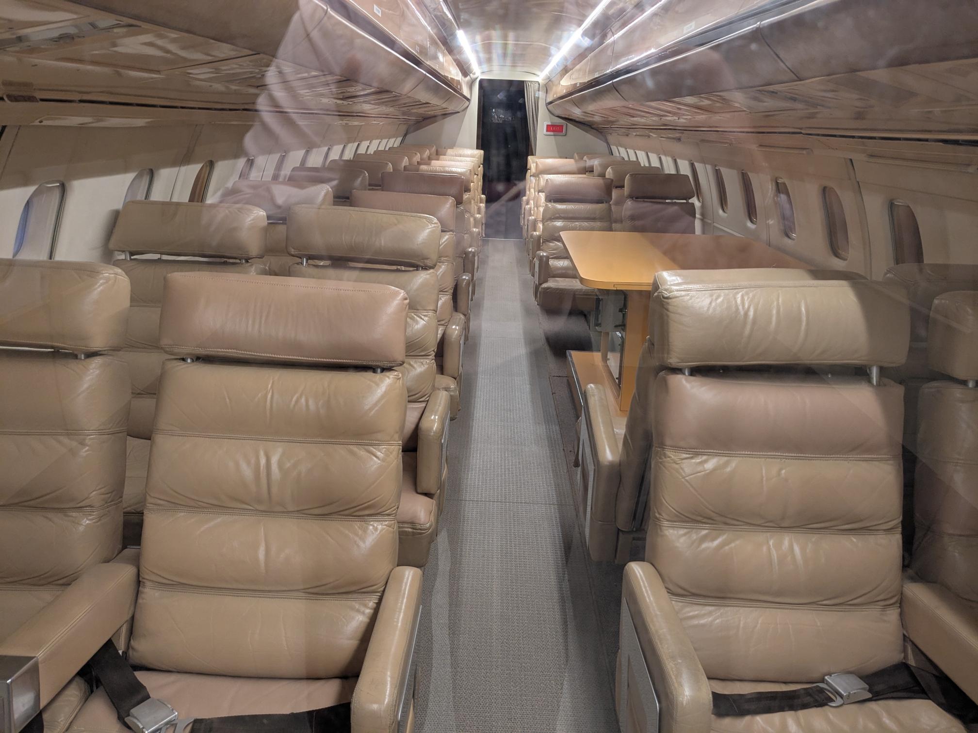 rangées de sièges en cuir beige (2+2), une allée centrale, dans un avion. il y a un espace avec une table derrière les premiers sièges à droite
