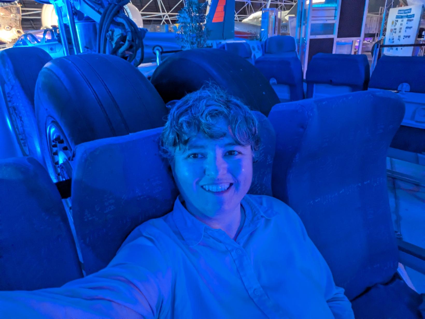 Selfie sur un siège d'avion. La teinte de la photo est bleue à cause des lumières d'ambiance. Juste derrière la rangée de sièges, on voit les roues d'un train d'atterrissage. cette personne n'est clairement pas dans un avion.